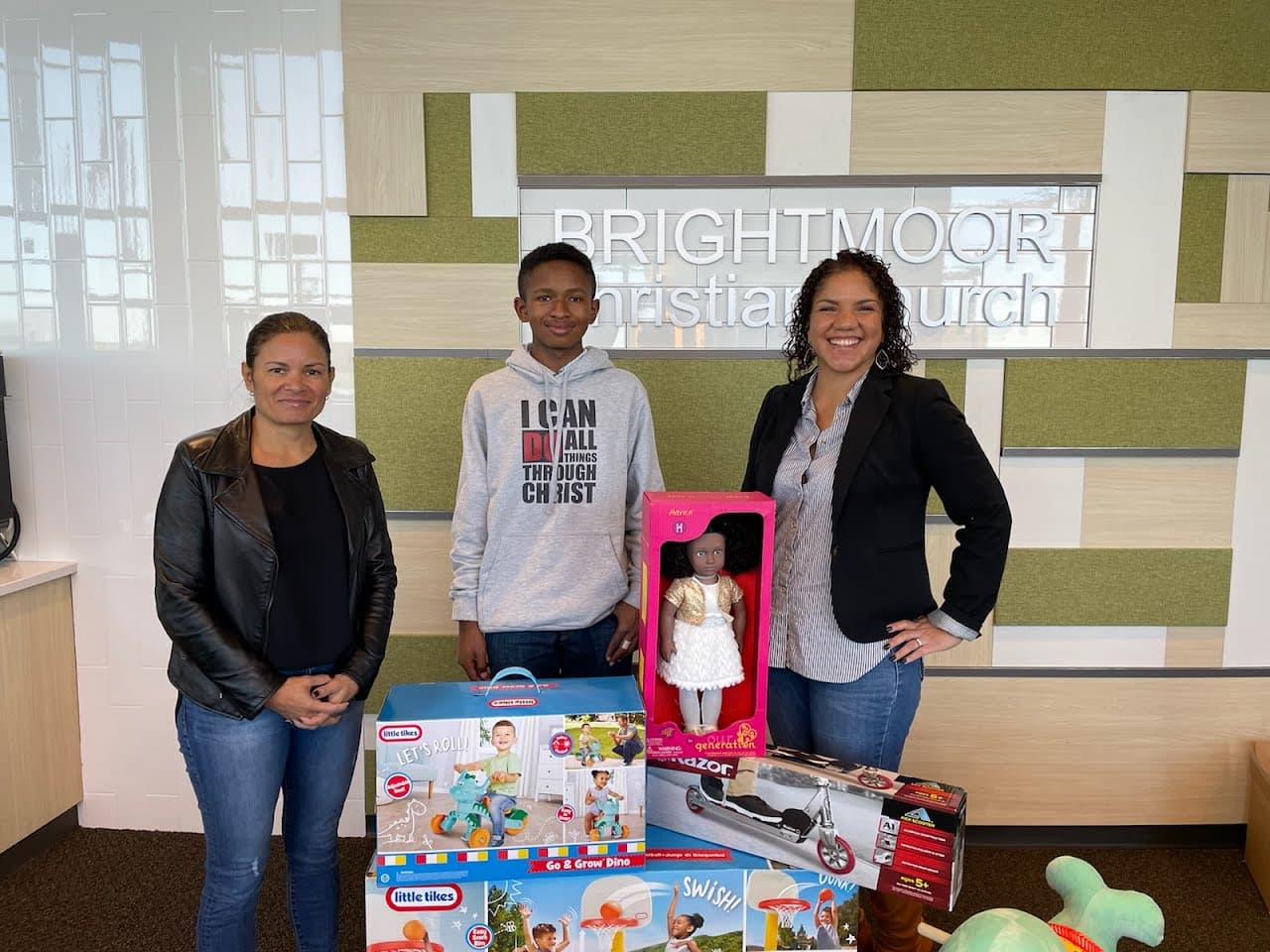 Three people posing with donated toys in front of a Brightmoor Christian Church sign.