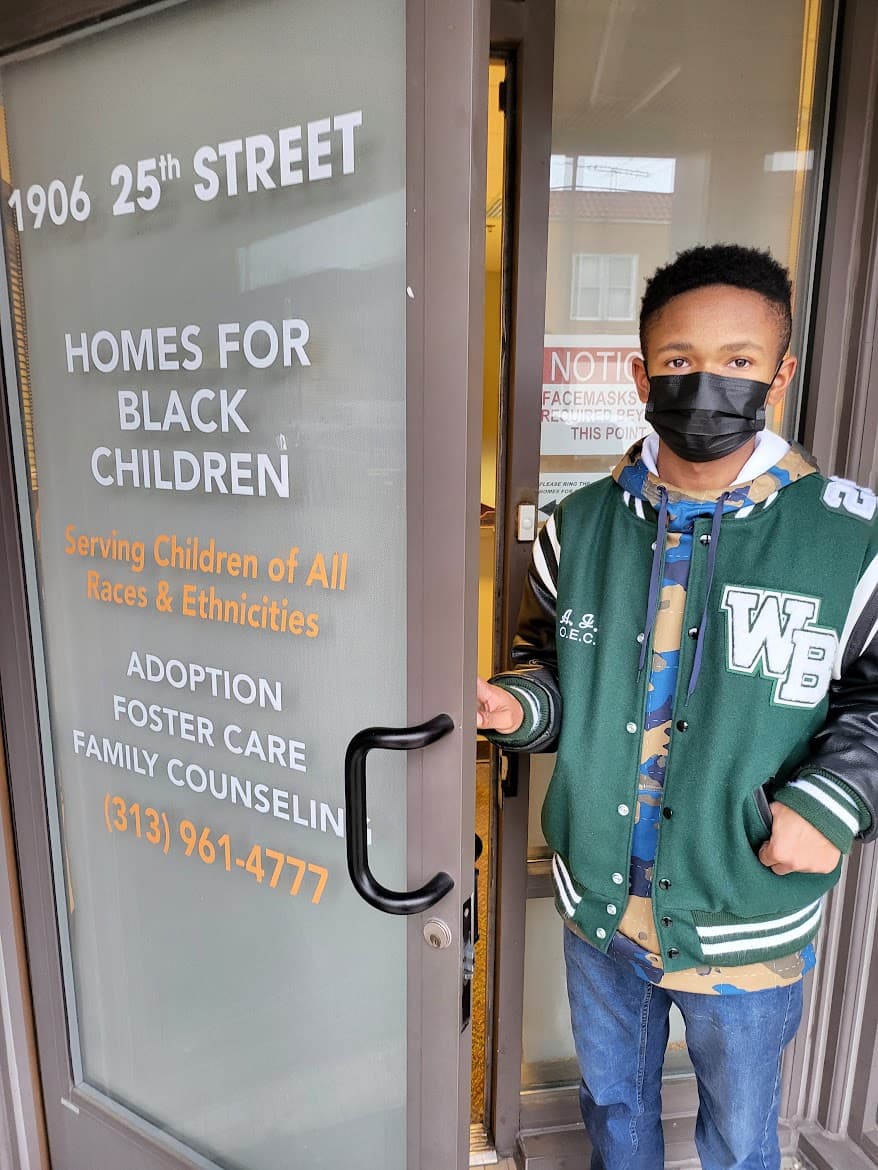 Masked young man in a green letterman jacket at the Homes for Black Children entrance.