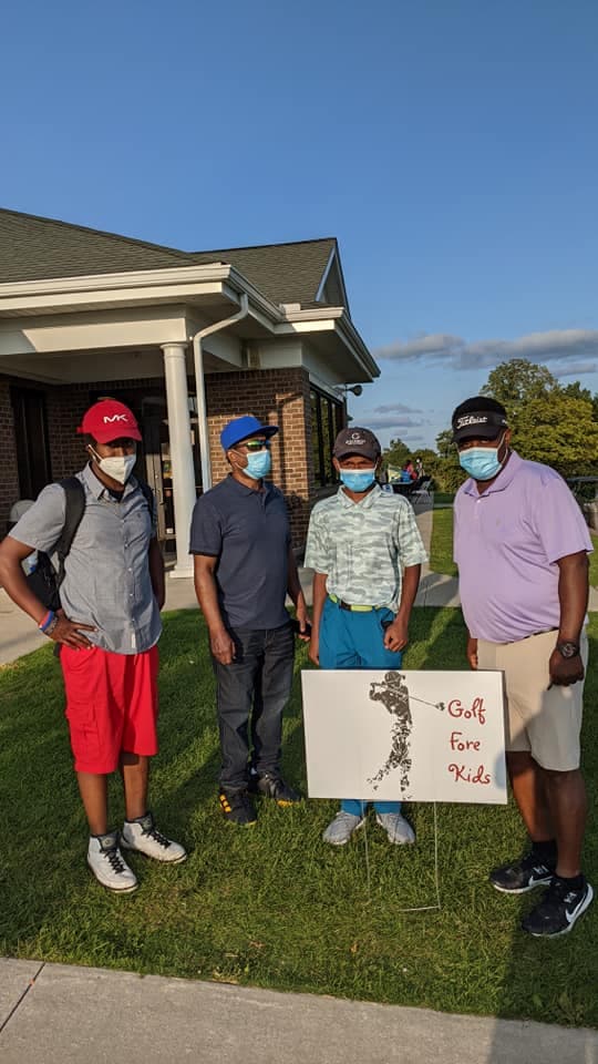 Four men wearing face masks and golf attire pose beside a Golf Fore Kids sign.