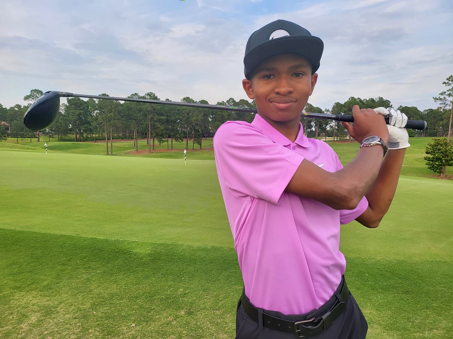 Young golfer in pink polo posing with a driver across his shoulders on a green.